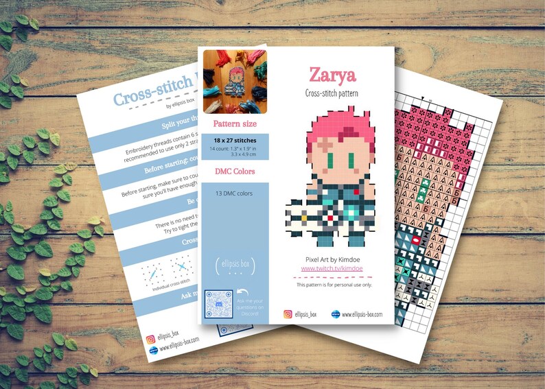 May include: Three sheets of paper with cross-stitch patterns on a wooden surface. The main sheet features a pixel art design of a character named Zarya, with a pink hairstyle. The pattern size is 18 x 27 stitches (4.6 x 6.9 cm), and 13 DMC colours are used.