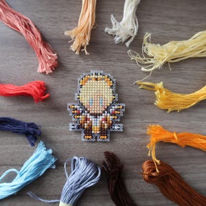 May include: A cross-stitch design of a figure with a yellow head and brown and gold body, surrounded by colorful embroidery floss in shades of red, orange, yellow, blue, and brown. The floss is arranged around the figure on a wooden surface.