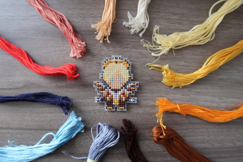 May include: A cross-stitch design of a figure with a yellow head and brown and gold body, surrounded by colorful embroidery floss in shades of red, orange, yellow, blue, and brown. The floss is arranged around the figure on a wooden surface.