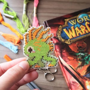 May include: A pixelated green creature with orange accents, possibly a video game character, is held in a hand. Embroidery floss in various colors and a "World of Warcraft" book are in the background. Scissors and an orange marker are also visible.