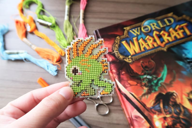 May include: A pixelated green creature with orange accents, possibly a video game character, is held in a hand. Embroidery floss in various colors and a "World of Warcraft" book are in the background. Scissors and an orange marker are also visible.