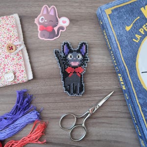 May include: A collection of craft supplies and accessories, including a floral patterned notebook, embroidery floss, scissors, and a cross-stitch cat design. A book with the title "Kiki La Petite Sorci&egrave;re" is also present.