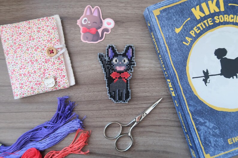 May include: A collection of craft supplies and accessories, including a floral patterned notebook, embroidery floss, scissors, and a cross-stitch cat design. A book with the title "Kiki La Petite Sorci&egrave;re" is also present.