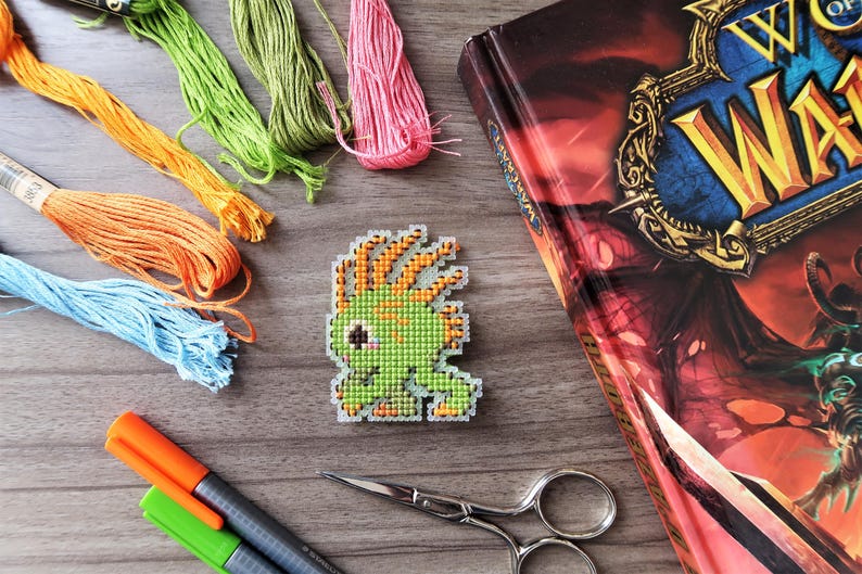 May include: A cross-stitch design of a green creature with orange hair, next to a book with a gold title. Colorful embroidery floss in orange, blue, green, and pink, along with scissors and markers, are also visible.