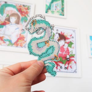 Op de afbeelding: Een kruissteekdraak in tinten van teal, wit en paars wordt getoond voor ingelijste anime-kunst. De draak heeft een gedetailleerd, gepixeld ontwerp. De kunstwerken op de achtergrond bevatten kleurrijke illustraties.