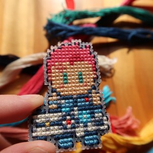 May include: A small, pixelated cross-stitch figure with red hair and blue clothing. The figure is held in a hand, with colourful embroidery threads in the background. This handmade craft project is a unique item.