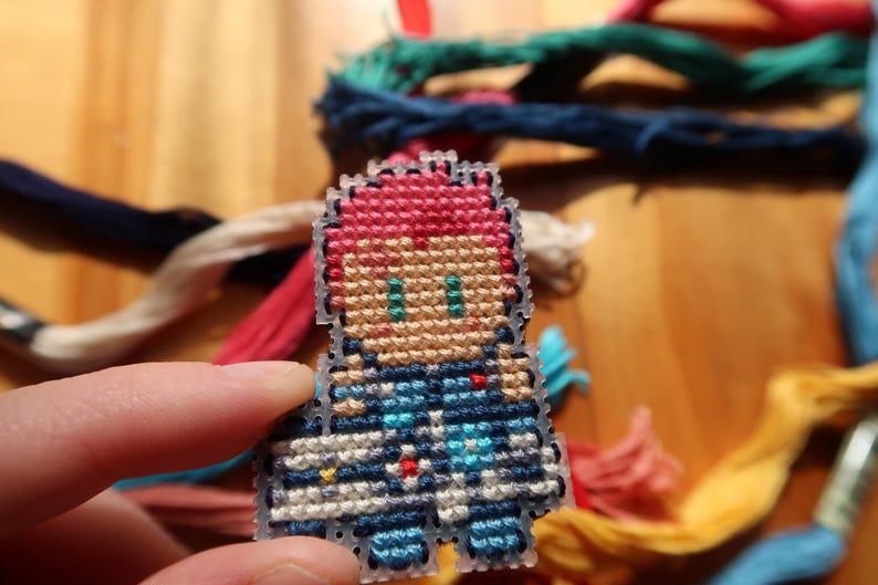 May include: A small, pixelated cross-stitch figure with red hair and blue clothing. The figure is held in a hand, with colourful embroidery threads in the background. This handmade craft project is a unique item.