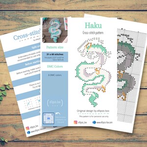 Op de afbeelding: Een verzameling kruissteekpatroonvellen op een houten oppervlak. Het hoofdvak toont een drakenontwerp met de naam "Haku" met een patroongrootte van 31 x 60 steken. Extra vellen geven instructies en kleurendetails. Het ontwerp gebruikt 6 DMC-kleuren.