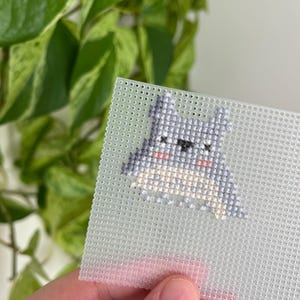 May include: A small, cross-stitched design of a cartoon character with gray, white, and pink details. The design is on a clear plastic canvas with a grid of small holes. The background includes green leafy foliage.