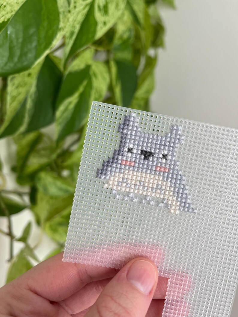 May include: A small, cross-stitched design of a cartoon character with gray, white, and pink details. The design is on a clear plastic canvas with a grid of small holes. The background includes green leafy foliage.