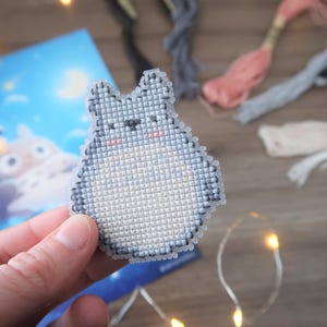 May include: A cross-stitch of a gray Totoro character with a white belly and pink cheeks. The craft is held in front of a blue illustration and colorful embroidery floss. The background includes fairy lights.