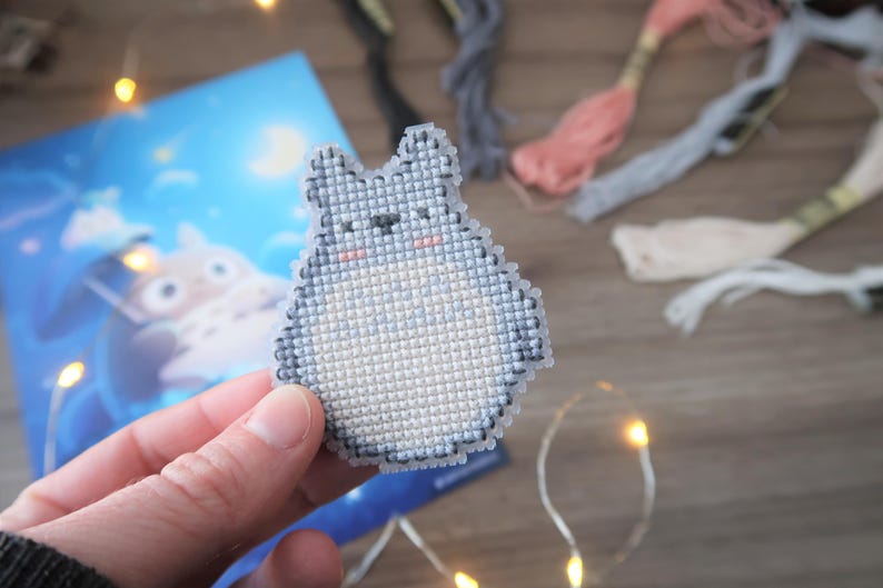 May include: A cross-stitch of a gray Totoro character with a white belly and pink cheeks. The craft is held in front of a blue illustration and colorful embroidery floss. The background includes fairy lights.