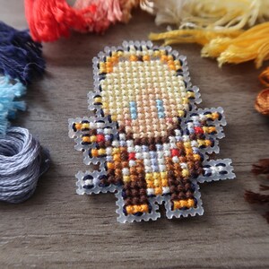 May include: A cross-stitch design of a pixelated character with blonde hair, blue eyes, and a brown and orange outfit. The design is surrounded by various colors of embroidery floss, including blue, red, yellow, and brown. The craft supplies are arranged on a wooden surface.