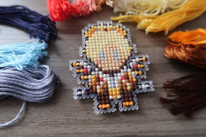 May include: A cross-stitch design of a pixelated character with blonde hair, blue eyes, and a brown and orange outfit. The design is surrounded by various colors of embroidery floss, including blue, red, yellow, and brown. The craft supplies are arranged on a wooden surface.