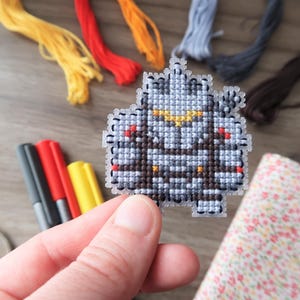 May include: A hand holds a cross-stitch project featuring a pixelated design in shades of gray, brown, and yellow. Embroidery floss in yellow, red, orange, and blue are in the background, along with markers and scissors. The project is a craft item.