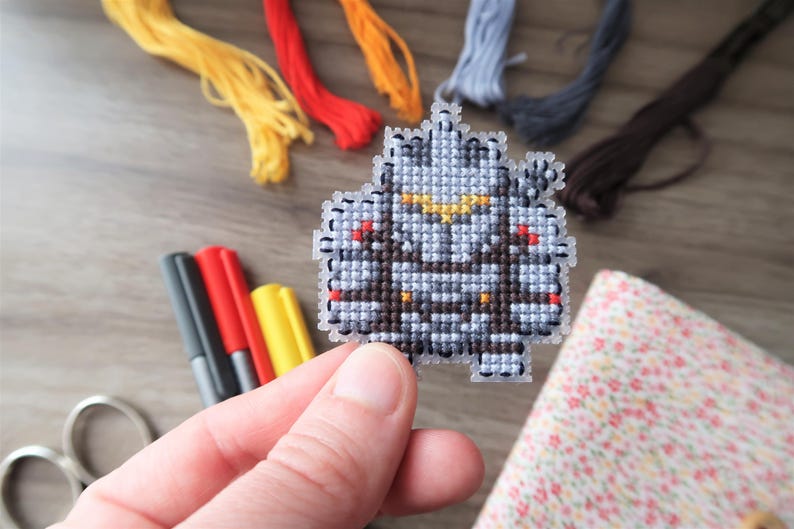 May include: A hand holds a cross-stitch project featuring a pixelated design in shades of gray, brown, and yellow. Embroidery floss in yellow, red, orange, and blue are in the background, along with markers and scissors. The project is a craft item.