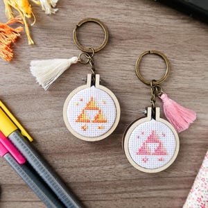 May include: Two keyrings with cross-stitch designs. One features a yellow and orange triangle design, the other a pink triangle. Both have wooden hoops, bronze-coloured key rings, and decorative tassels. Other craft supplies are also visible.