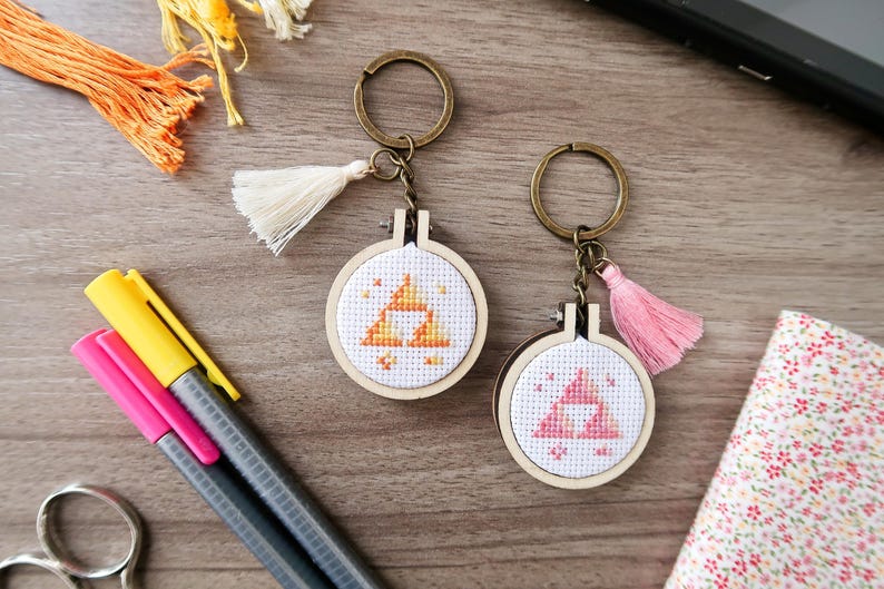 May include: Two keyrings with cross-stitch designs. One features a yellow and orange triangle design, the other a pink triangle. Both have wooden hoops, bronze-coloured key rings, and decorative tassels. Other craft supplies are also visible.