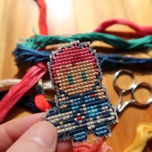 May include: A cross-stitch figure in pixel art style, featuring red hair and blue clothing. The figure is set against a backdrop of colourful embroidery threads and small scissors on a wooden surface.