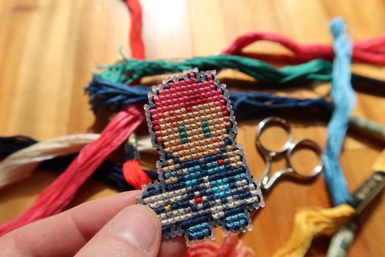 May include: A cross-stitch figure in pixel art style, featuring red hair and blue clothing. The figure is set against a backdrop of colourful embroidery threads and small scissors on a wooden surface.