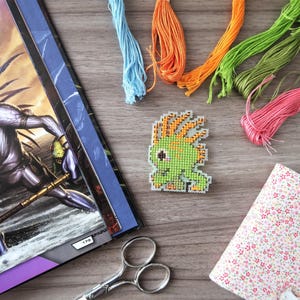 May include: A cross-stitch project featuring a green and orange pixelated creature, surrounded by colorful embroidery floss in blue, orange, green, and pink. A floral patterned notebook and scissors are also present, suggesting a crafting theme.