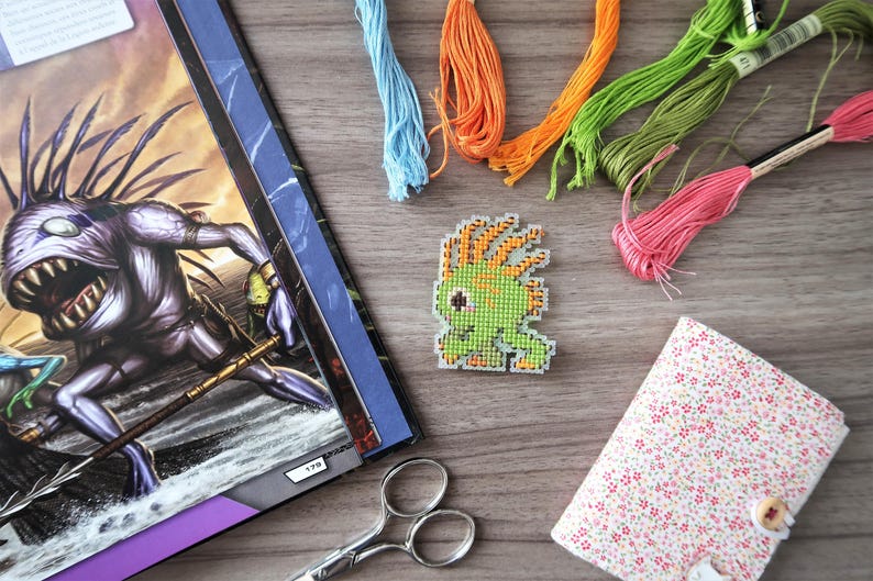 May include: A cross-stitch project featuring a green and orange pixelated creature, surrounded by colorful embroidery floss in blue, orange, green, and pink. A floral patterned notebook and scissors are also present, suggesting a crafting theme.