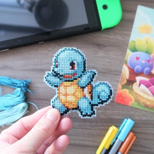 May include: A handmade cross-stitch of a blue Squirtle character with an orange shell. The image also shows a Nintendo Switch, a colorful illustration of other Pokemon characters, blue embroidery floss, and markers.