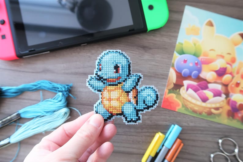 May include: A handmade cross-stitch of a blue Squirtle character with an orange shell. The image also shows a Nintendo Switch, a colorful illustration of other Pokemon characters, blue embroidery floss, and markers.