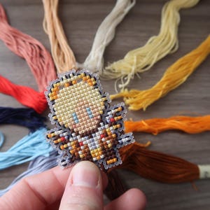 May include: A cross-stitch design of a small figure with blonde hair and blue eyes, held in a hand. The figure is surrounded by colorful embroidery floss in shades of red, pink, yellow, and blue.