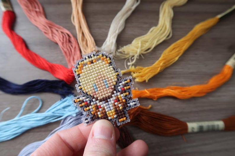 May include: A cross-stitch design of a small figure with blonde hair and blue eyes, held in a hand. The figure is surrounded by colorful embroidery floss in shades of red, pink, yellow, and blue.