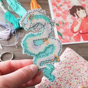 Op de afbeelding: Een hand houdt een kruissteekdraak in tinten blauw, wit en goud vast. Borduurgaren in verschillende kleuren en een print van een anime-personage staan op de achtergrond. Een kleine schaar en bloemenstof zijn ook zichtbaar.