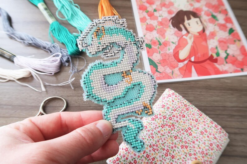 Op de afbeelding: Een hand houdt een kruissteekdraak in tinten blauw, wit en goud vast. Borduurgaren in verschillende kleuren en een print van een anime-personage staan op de achtergrond. Een kleine schaar en bloemenstof zijn ook zichtbaar.