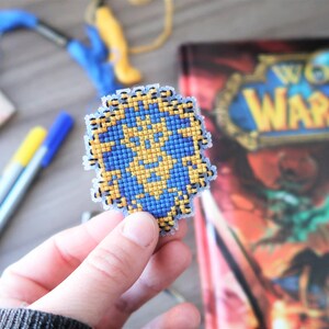 May include: A handmade, pixelated shield emblem in blue and gold, held in a person's hand. The shield is in front of a World of Warcraft book. Crafting supplies, including pens and embroidery floss, are in the background.