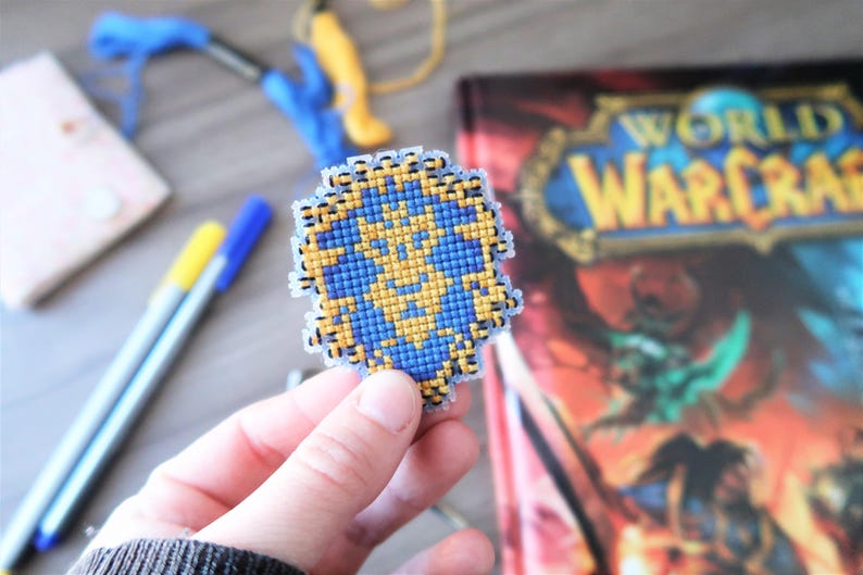 May include: A handmade, pixelated shield emblem in blue and gold, held in a person's hand. The shield is in front of a World of Warcraft book. Crafting supplies, including pens and embroidery floss, are in the background.