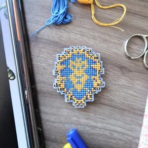 May include: A handmade cross-stitch design in blue and yellow, featuring a geometric pattern. The craft project is surrounded by blue and yellow thread, scissors, and a floral fabric swatch, all on a wooden surface.