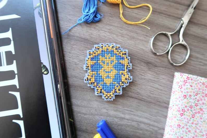 May include: A handmade cross-stitch design in blue and yellow, featuring a geometric pattern. The craft project is surrounded by blue and yellow thread, scissors, and a floral fabric swatch, all on a wooden surface.