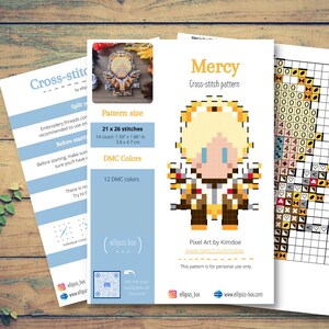 May include: Cross-stitch pattern featuring a pixel art design of a character named Mercy. The pattern size is 1.50 x 1.86 inches, with 21 x 26 stitches. The design uses 12 DMC colors. The image is on a wooden surface.