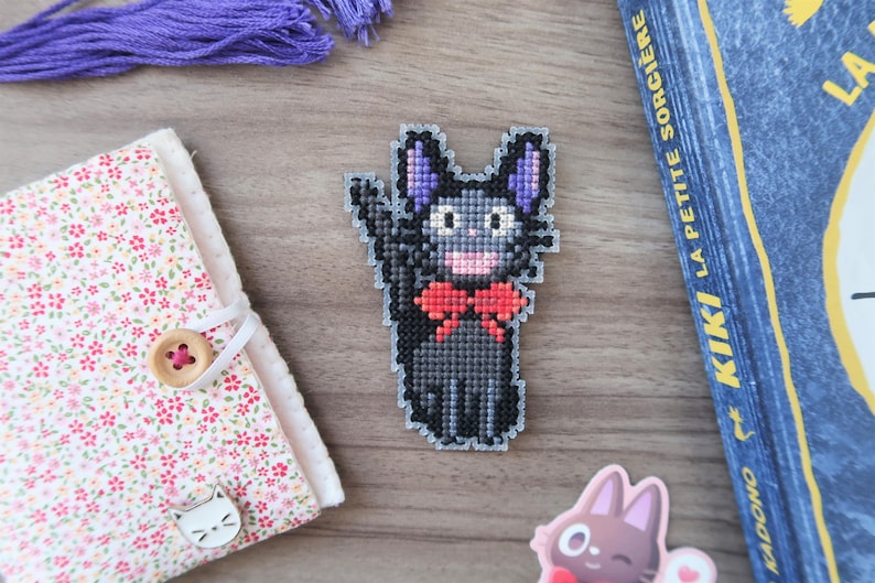 May include: A cross-stitch cat with a red bow tie, purple ears, and a waving paw. The cat is on a wooden surface, next to a floral notebook, purple thread, and a book titled "Kiki La Petite Sorciere".