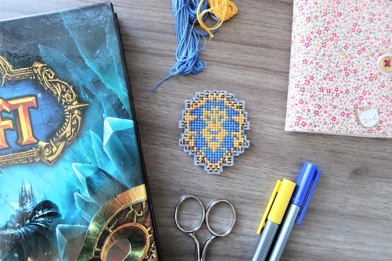 May include: A cross-stitch emblem in blue and yellow, featuring a detailed design. The emblem is surrounded by craft supplies, including blue and yellow thread, scissors, and markers. A book with a blue cover and a floral-patterned notebook are also present.