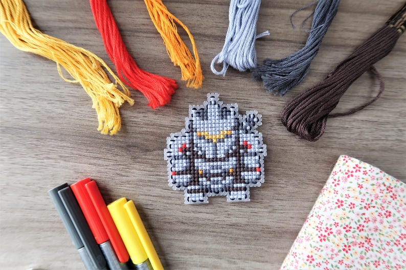 May include: A cross-stitch project featuring a pixelated robot design. The image includes colorful embroidery floss in yellow, red, orange, gray, and brown, along with a set of markers and a floral fabric swatch. The craft supplies are arranged on a wooden surface.