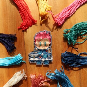 May include: A cross-stitch character with pink hair and blue clothing is surrounded by colourful embroidery floss in shades of red, yellow, pink, green, blue, and white. The craft supplies are arranged on a wooden surface.