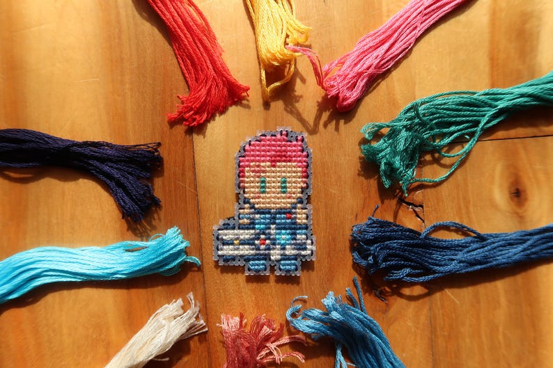 May include: A cross-stitch character with pink hair and blue clothing is surrounded by colourful embroidery floss in shades of red, yellow, pink, green, blue, and white. The craft supplies are arranged on a wooden surface.