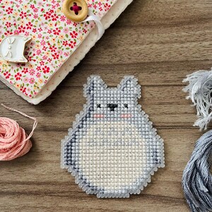 May include: A handmade cross-stitch design of a cartoon character in shades of gray and white, with pink cheeks. The design is placed on a wooden surface, next to a floral fabric-covered notebook with a wooden button closure.