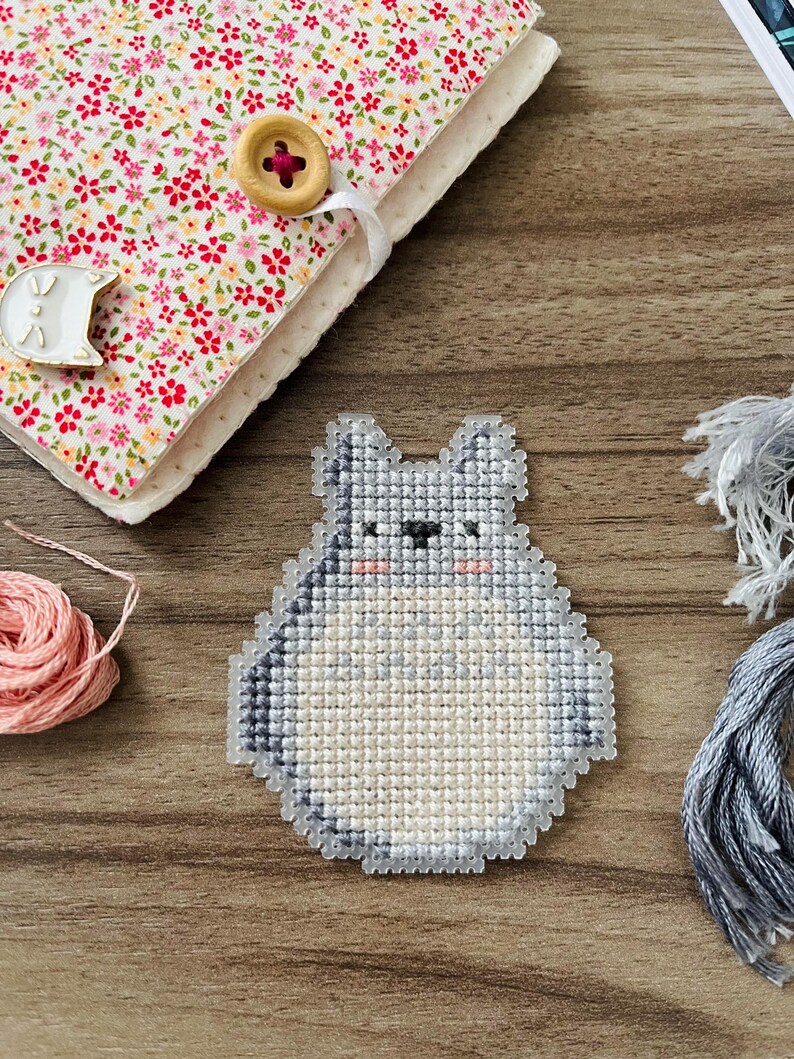 May include: A handmade cross-stitch design of a cartoon character in shades of gray and white, with pink cheeks. The design is placed on a wooden surface, next to a floral fabric-covered notebook with a wooden button closure.