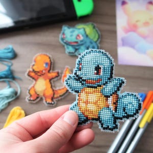May include: Cross-stitch Pokemon characters, including Squirtle, Charmander, and Bulbasaur. The Squirtle is held in a hand. Other craft supplies, including embroidery floss and pens, are also visible.