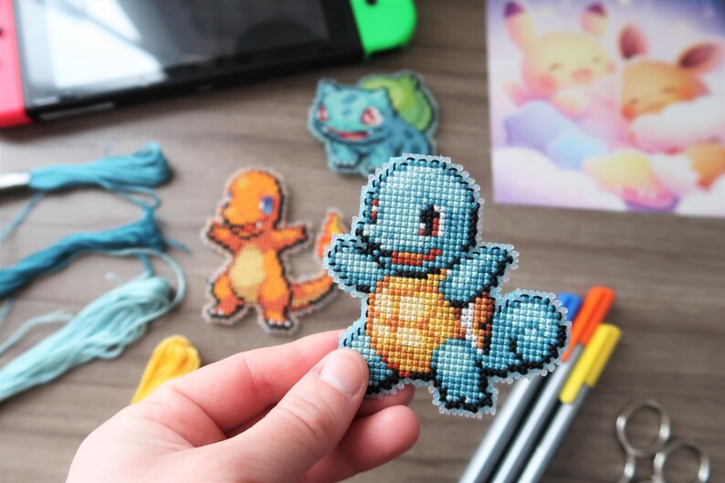 May include: Cross-stitch Pokemon characters, including Squirtle, Charmander, and Bulbasaur. The Squirtle is held in a hand. Other craft supplies, including embroidery floss and pens, are also visible.