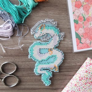 Op de afbeelding: Een kruissteekdraak in tinten blauw, wit en paars, met gouden accenten. De draak is omgeven door borduurgaren in verschillende kleuren, een schaar en een stof met bloemenprint. Er is ook een afdruk van een anime-personage.