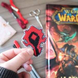 May include: A handmade, red and black cross-stitch emblem, held in a hand, featuring a stylized design. The emblem is likely a logo or symbol. A World of Warcraft guide book is in the background, along with craft supplies.
