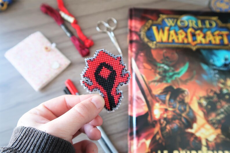 May include: A handmade, red and black cross-stitch emblem, held in a hand, featuring a stylized design. The emblem is likely a logo or symbol. A World of Warcraft guide book is in the background, along with craft supplies.