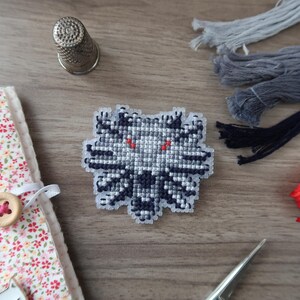 May include: A cross-stitch patch featuring a wolf's head design with red eyes, in shades of gray and black. The patch is surrounded by embroidery floss in gray, black, and red, a thimble, a needle, and a floral fabric-covered notebook.